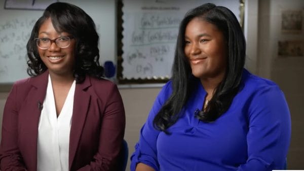 Two students Calcea Johnson and Ne'Kiya Jackson interview in classroom for the news. The girl on the left is wearing a white shirt with a burgundy blazer on top and glasses with short curled hair and the one on the right is wearing long straight hair with a blue blouse.